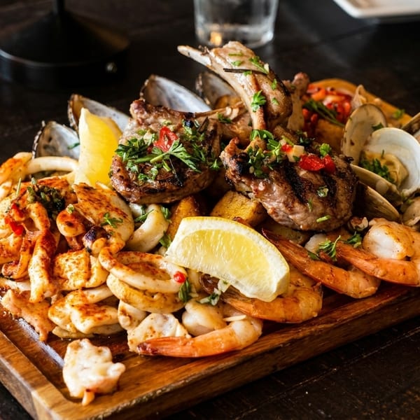 A large wooden platter overflowing with grilled lamb chops, shrimp, calamari, clams, and scallops.