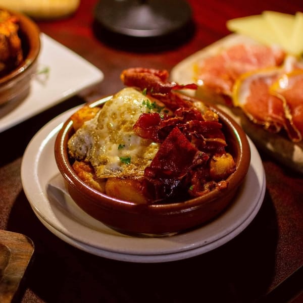 Baked casserole topped with melted cheese, crispy bacon, and herbs, served in a brown dish alongside bread and sliced meat.