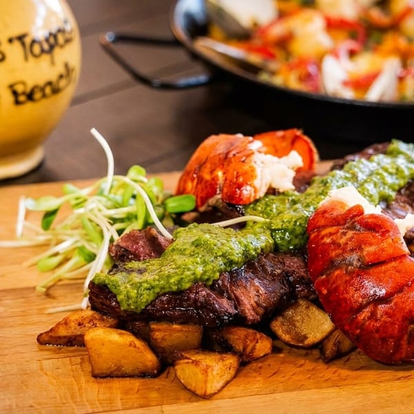 Steak and lobster tails with green sauce and potatoes on a wooden board at Papa's Tapas.